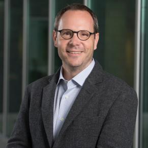 A man in a grey jacket and blue shirt wearing spectacles smiles at the camera.