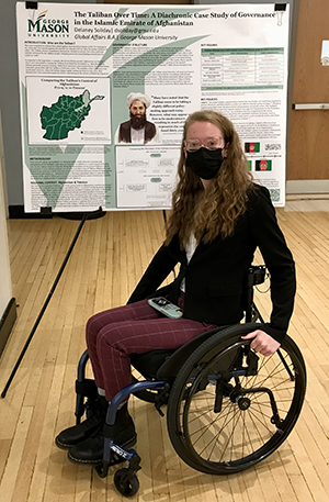 A young woman in eye glasses and a black mask sits in a wheelchair in front of a poster she created about the Taliban.