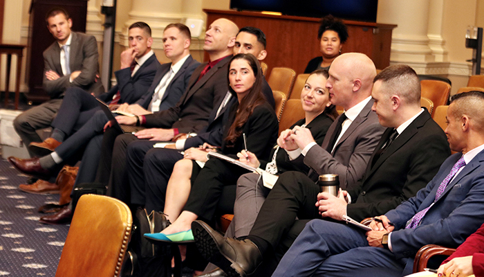 Men in suits and women in dresses sit in leather chairs in a row.