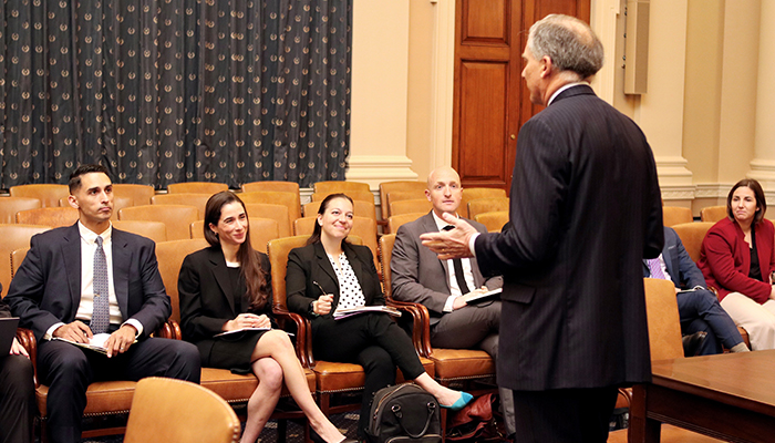Six men and women sit in brown leather chairs as a man in a dark suit speaks to them.