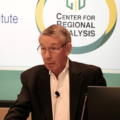 A man in a suit stands at a podium with a slide on a screen behind him.