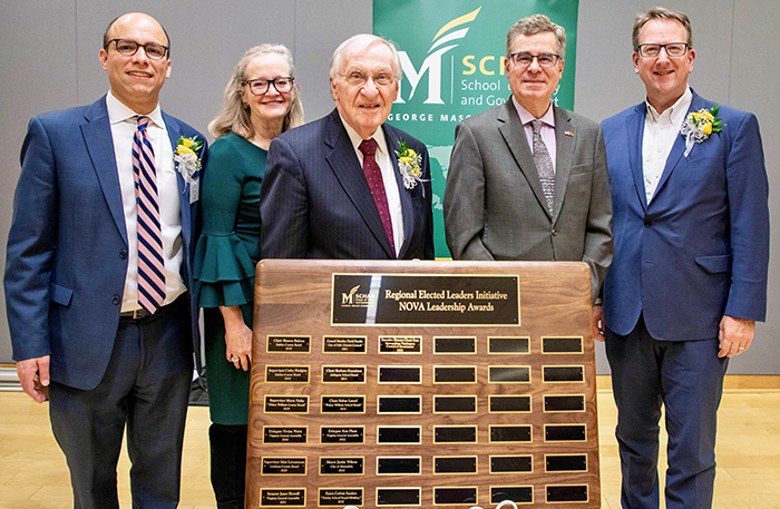 Five people dressed for success stand in front of a wooden plaque.