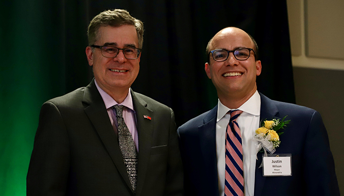 Two men in suits, one with a corsage, pose in front of a green curtain.