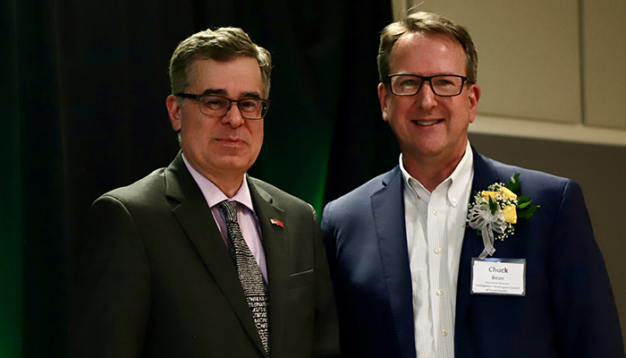 Two men in suits, one with a corsage, pose in front of a green curtain.