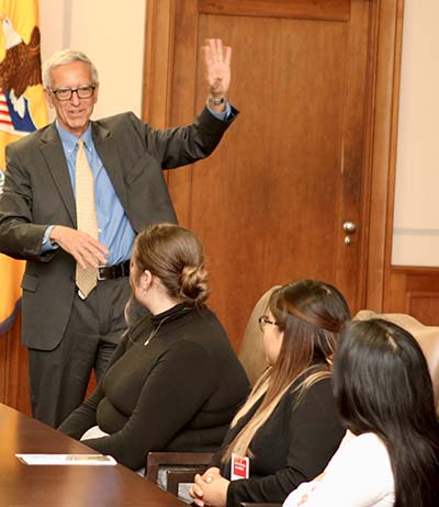 A man in a suit gestures at onlookers sitting at a table.