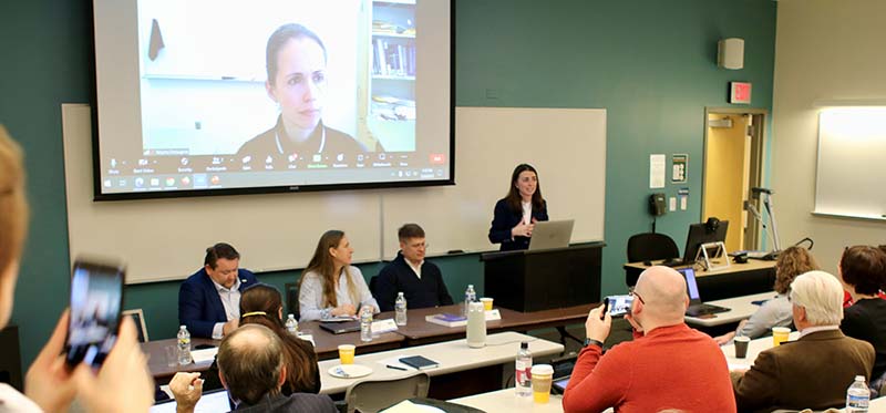 Three people sit at a table while a woman at a podium gestures in a classroom with people watching.