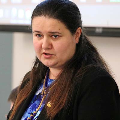 A woman with brown hair in a blue top with black jacket makes a comment.