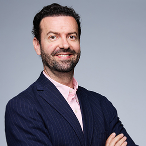 A dark-haired man in a blue jacket and pink shirt looks at the camera.