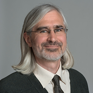 A man with long silver hair and a short beard stares at the camera.