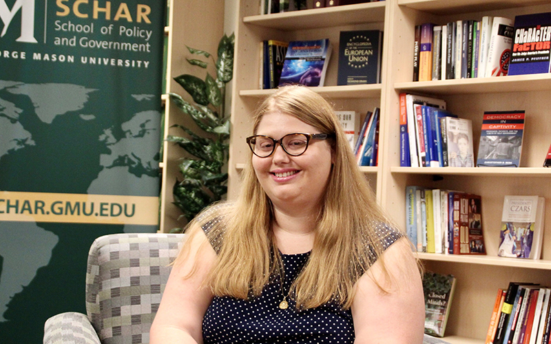 A woman with long blonde hair sits on a checked couch in front of books and a Schar School banner.