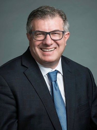 A man in a black suit jacket, white shirt, and blue tie smiles at the camera.
