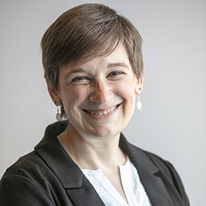 A woman in a dark jacket and light blouse with short brown hair smiles at the camera.