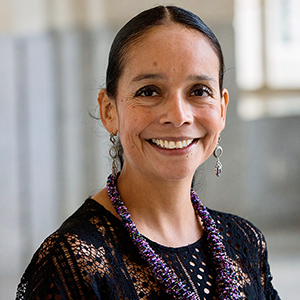 A woman with her hair pulled back and wearing a blue knit dress and a purple necklace smiles at the camera.