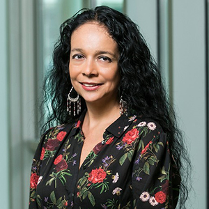 A woman with long black hair and in a black floral top looks at the camera.