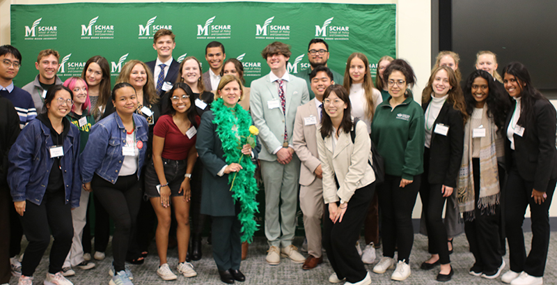 Twenty-four students pose in front of a green Schar School sign with a woman in the center as she wears a green boa and holds a yellow flower.