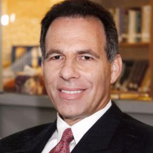 Jack Goldstone stands smiling while wearing a black suit, white collared shirt, and red tie