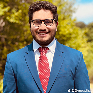 Maaz Abbasi standing outside and smiling while wearing a blue suit, white shirt, and red tie
