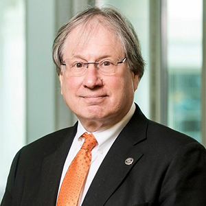 A man in eyeglasses, a black jacket, orange necktie, and thinning hair stands in front of a greenish background.