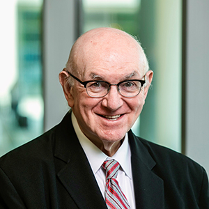 A man with very little hair, eyeglasses, and a big smile poses in front of a green backdrop.