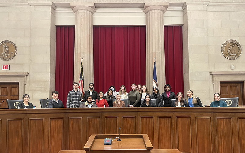 A group of students sit and stand behind a wooden dais and in front of a red curtain and two large pillars.