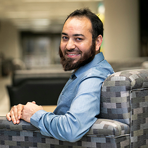 A man with a beard and wearing a blue shirt sits in a chair facing the camera.