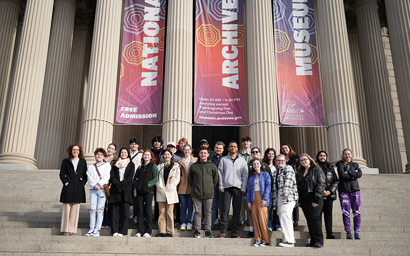 A group of students stands on stairs in front of a columned building with three banners hanging.