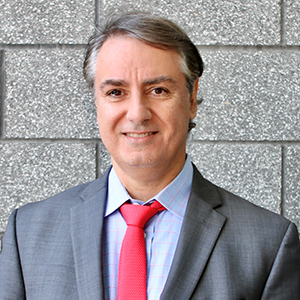 A man in a gray suit, blue shirt, and red tie with gray temples gazes at the camera.