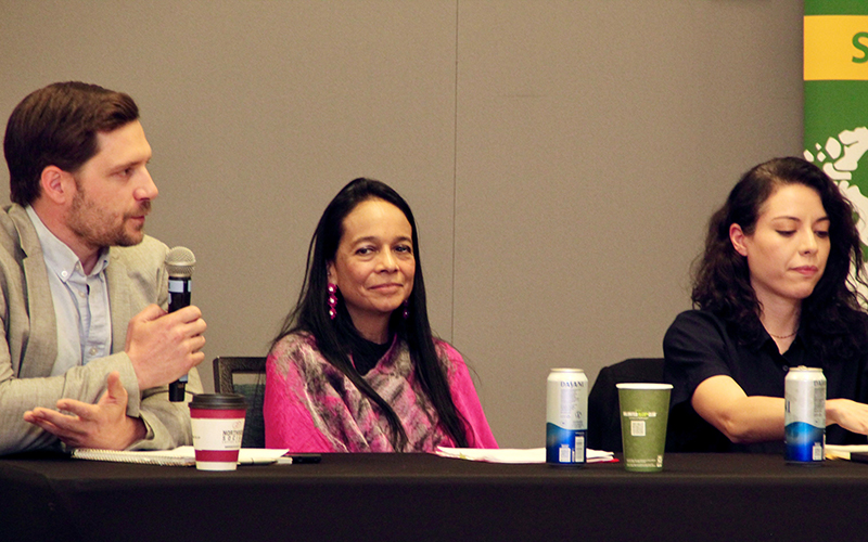 A man in a beige suit and holding a microphone sits at a table with a woman in pink and a woman in black.