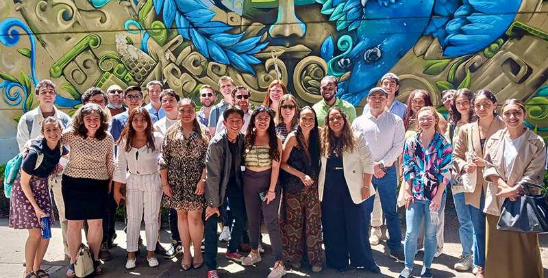 A large group of college students pose in front of a brightly colored mural.