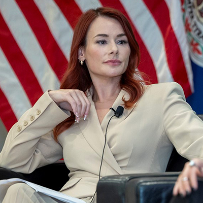 A woman with bright red hair in a cream-colored pantsuit sits in front of an American flag.