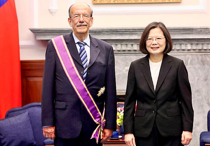 A man in a blue suit with a purple and gold sash across his chest stands next to a woman in a dark suit as they face the camera.