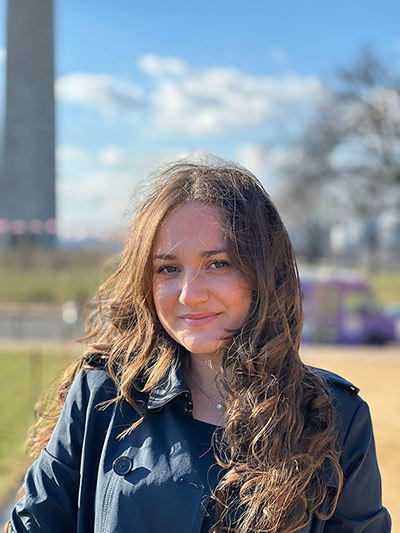 A young woman with long brown hair smiles and stands in front of the Washington Monument.