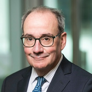 A man in eyeglasses and a dark jacket with a blue tie looks at the camera.