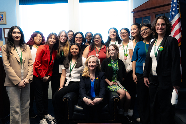 Several young women smile at the camera as they pose around a blond woman in a black jacket sitting on a black leather chair.