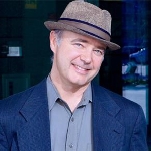 A man in a blue jacket and gray shirt wearing a hat smiles at the camera.
