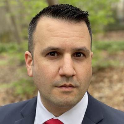 Lee Roberts stands outside smiling while wearing a gray suit, white collared shirt, and red tie