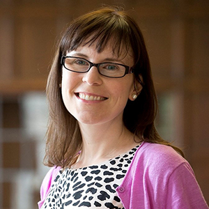 Catherine Maclean stands inside smiling while wearing a pink sweater