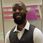 A man with a beard wearing a gray vest and white shirt stands and smiles