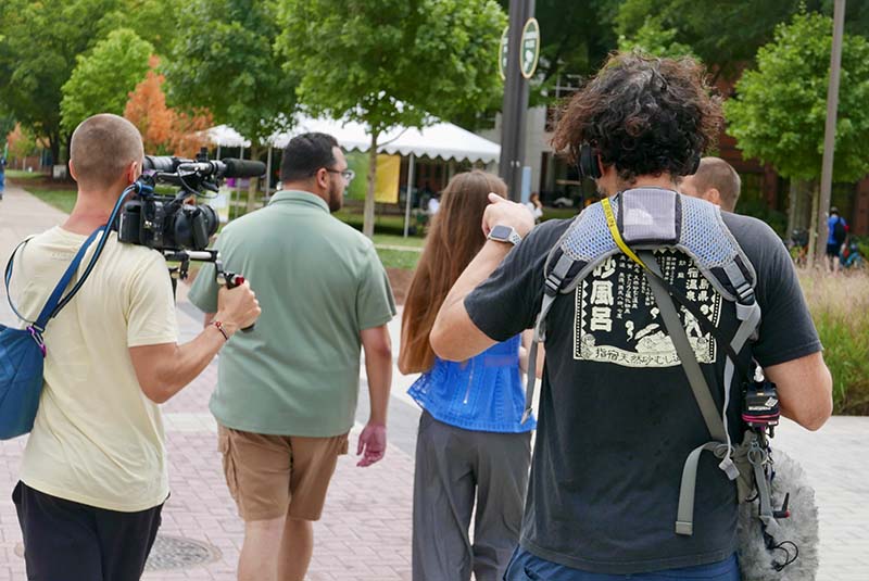 A man carrying a video camera walks behind a man in a green shirt and a woman in a blue top as another man in a black tee shirt follows them.