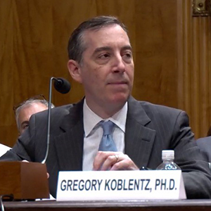 A man in a gray suit and blue tie sits at a table with a microphone.