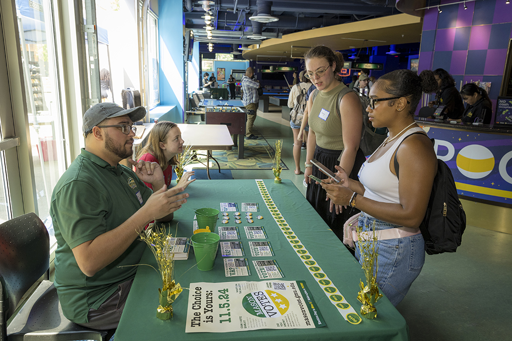 A young man wearing a baseball cap and a young woman sit at a table and talk to two women who are standing on the other side.