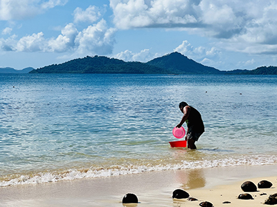 A man fills a red container with ocean water on a beach with mountains in the background.