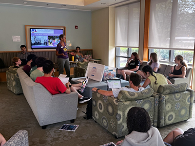 A woman stands and gestures with her hands in the center of a lounge with students sitting on chairs and couches.