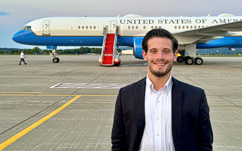 A man with dark hair, a short beard, a white shirt, and a dark jacket stands in front of a jet labeled United States of America.