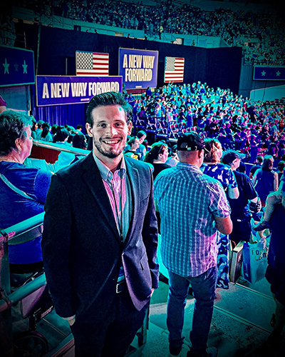 A young man in a dark suit stands amid a crowd in an arena during a presidential campaign event.