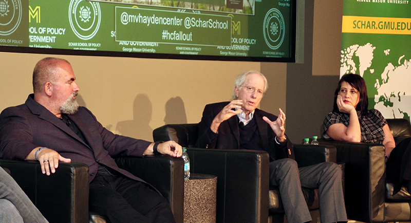Three people in chairs look at the man in the middle who gestures with his hands.