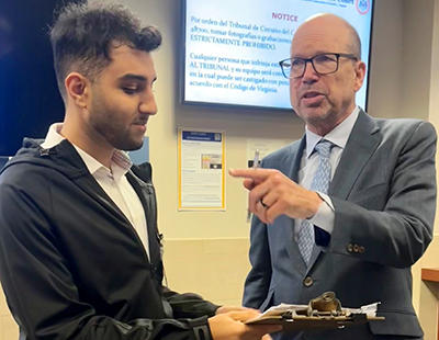 A man with dark hair and wearing a dark jacket and slacks holds a clipboard for a man in a blue suit and eyeglasses.