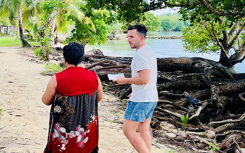 A man wearing a T-shirt, shorts, and flip-flops walks along a beach with a woman wearing a red and black dress.