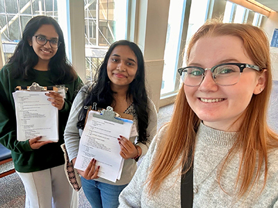 Three women, two of the showing white pages on clipboards, pose in a hallway.