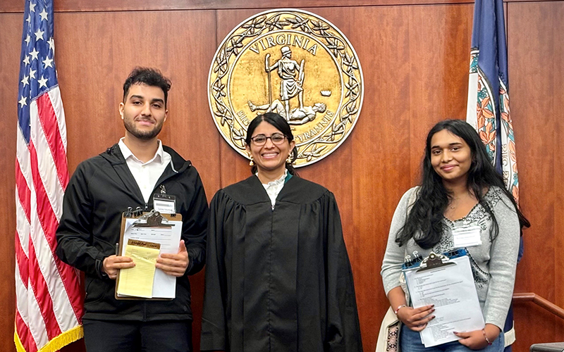 A man and a woman holding clipboards facing the camera stand on either side of a woman in a judge’s robe in a courtroom.